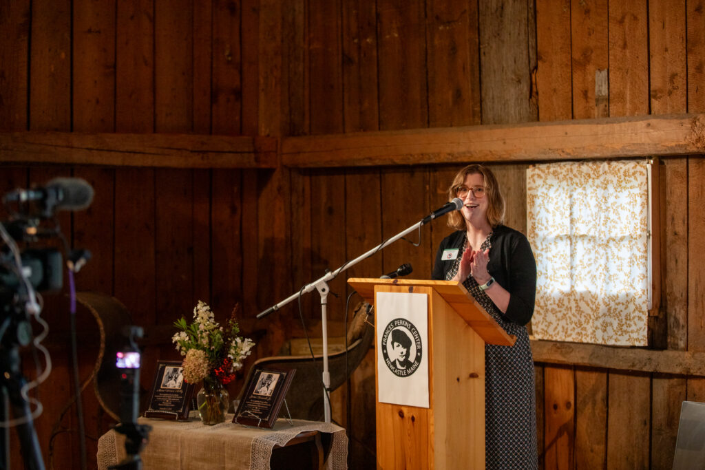 Woman standing at podium speaking into a microphone at the Frances Perkins National Monument inside a barn